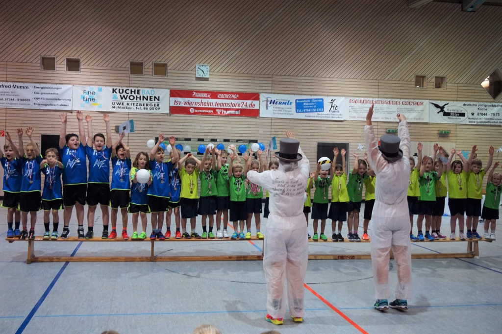 Kinder und zwei Erwachsene im weißen Kostüm auf einer Turnhallenbühne, halten Bälle hoch und klatschen gemeinsam bei einer Spielvorführung.