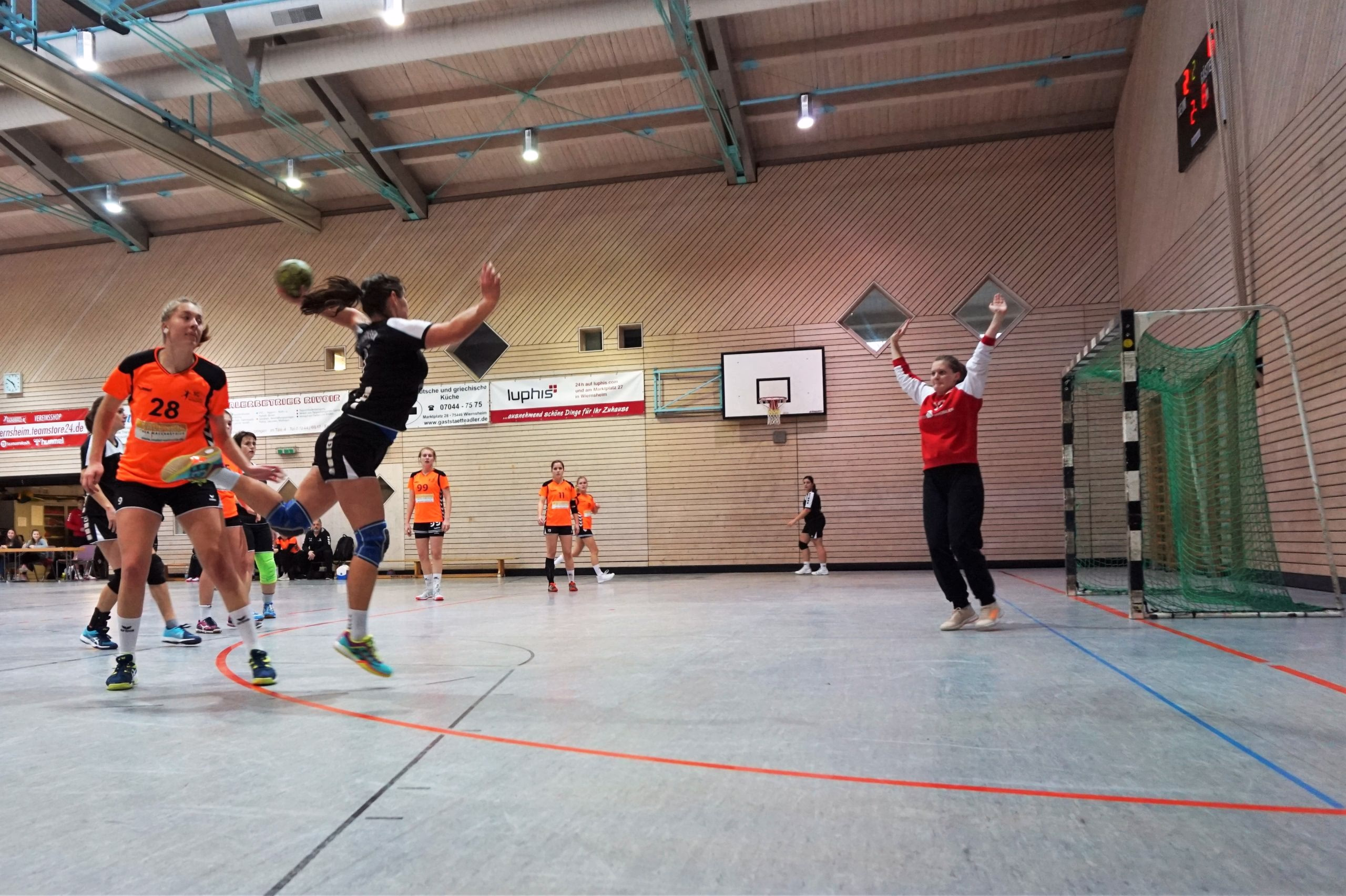 Handballspiel in der Halle: Spielerinnen springen und werfen, Schiedsrichterin hebt die Arme; rechts ist das Tor mit Netz zu sehen.