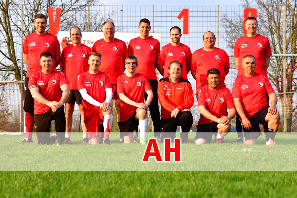 Mannschaftsfoto von Fußballspielern im roten Trikot auf dem Rasen, Hintergrund Trainingszaun; „1“ und „AH“ als Teaser/Platzhalter.