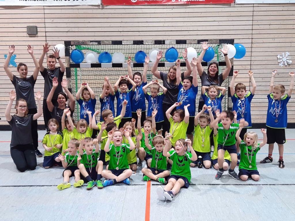 Gruppe von Kindern mit Medaillen auf dem Boden, recken die Hände und jubeln vor einem Tor mit Netz und großen blauen Ballons.