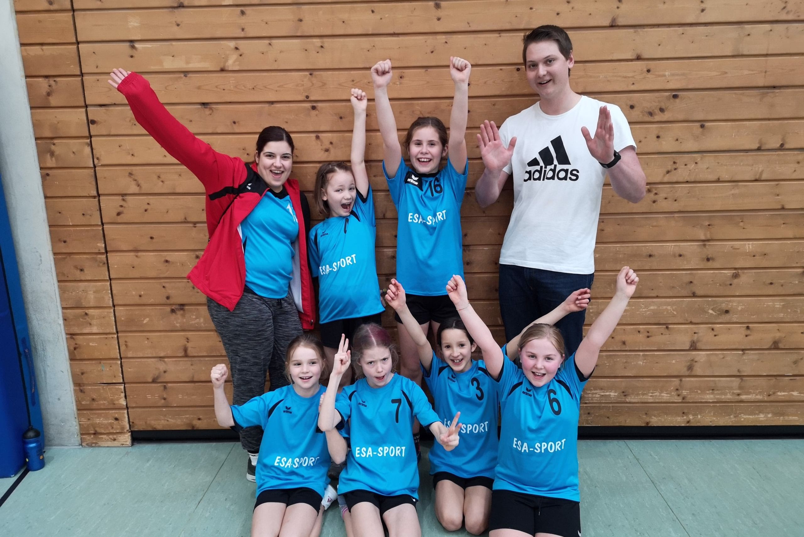 Kinderteam in Sportkleidung posiert vor Holzwand, hebt Fäuste und macht Siegergesten als Zeichen von Erfolg.