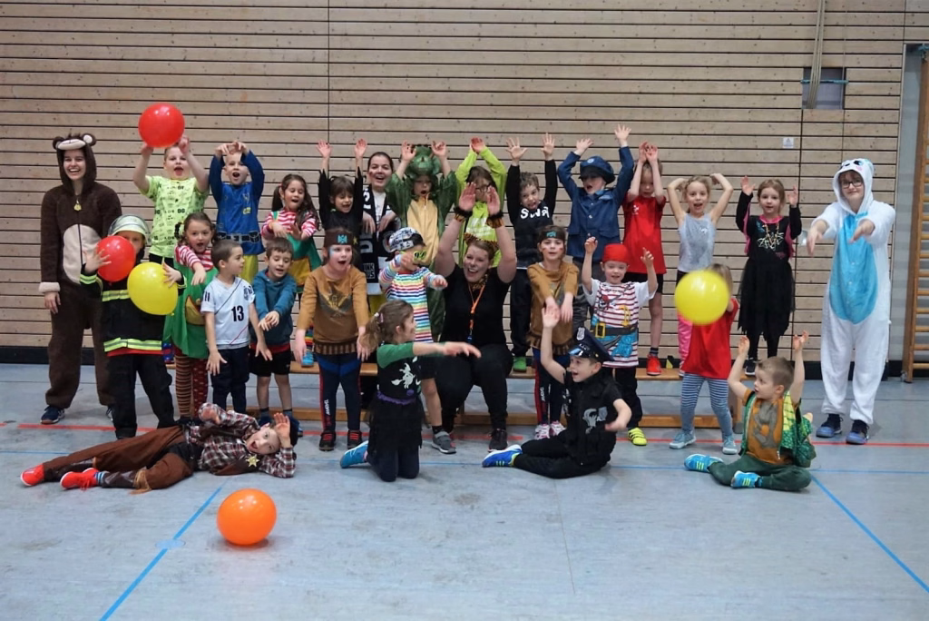 Gruppe von Kindern in bunten Kostümen in einer Turnhalle: Arme hoch, jubelnd, mit roten und gelben Luftballons zur Faschingsfeier.