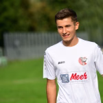 Junger Mann im weißen Trikot auf einem Fußballfeld, lächelt in die Kamera; Hintergrund mit unscharfen Zuschauergelände.