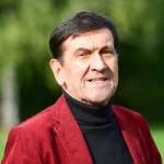 Lächelnder Mann im Porträt, Blick in die Kamera, rote Jacke über dunklem Rollkragenpullover; unscharfer grüner Hintergrund.