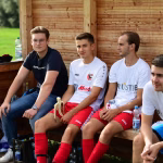 Vier Fußballer sitzen in einer Holzbankloge neben Getränken und Ausrüstung; Blicke nach vorne, während ein Spielakt abseits beobachtet wird.
