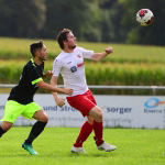 Zwei Fußballspieler kämpfen um den Ball in der Luft; Spieler im weißen Trikot zielt nach vorne, der Gegenspieler blockt beim Kopfballduell.