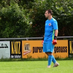 Fußballspieler in blauem Trikot und Torwarthandschuhen geht am Spielfeldrand vorbei, im Hintergrund eine Werbebande.