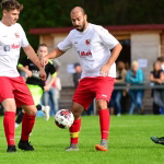 Drei Fußballspieler im Spiel: einer dribbelt mit dem Ball, andere laufen vorbei; Rasen und Zuschauer im Hintergrund.