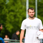 Porträt eines Mannes im Park mit weißem T‑Shirt und Handschuhen; hält eine Gabelung/Flasche und lächelt vor unscharfem Publikum.