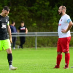 Zwei Fußballspieler auf dem Rasen, einer schaut nach unten, der andere steht mit Hand am Hüftbereich und beobachtet das Spielgeschehen.