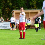 Fußballspieler auf dem Rasen beim Anstoß, im Vordergrund in Pose mit angewinkeltem Arm; weitere Spieler im Hintergrund bei einem Spiel.