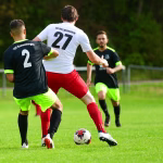 Zwei Fußballspieler jagen den Ball auf dem Rasen; ein Spieler mit Rückennummer führt den Lauf an, im Hintergrund ein weiterer Gegenspieler.