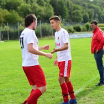 Zwei Fußballspieler im Trainingsfeld geben sich die Hand, während ein Trainer im roten Oberteil daneben zusieht; Sportlicher Moment vor dem Spiel.