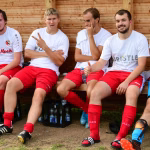 Fünf Fußballer sitzen in roten Trikots auf einer Holzbank, lehnen an der Wand und blicken in die Kamera; Team-Porträt.