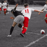 Zwei Fußballspieler prallen im Zweikampf auf nassem Rasen, ein Spieler rutscht nach vorn und setzt zum Torschuss an, Ball im Vordergrund.