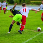 Zwei Fußballspieler prallen am Ball zusammen, einer in Neonorange deckt den Gegenspieler am Boden ab. Spielaktion im Fokus.