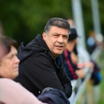 Mann in schwarzer Jacke im Gespräch, Blick zur Seite; im Vordergrund und Hintergrund unscharfe Zuhörer vor grünem Hintergrund.