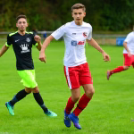 Zwei Fußballspieler rennen auf dem Rasen im Spiel, einer in weiß-roter Ausrüstung, der andere in schwarz-grün, angespannt im Zweikampf.