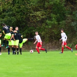 Fußballspiel auf dem Rasen: mehrere Spieler in Aktion, ein Spieler springt über den Ball, während andere abwehrend und angreifend positioniert sind.