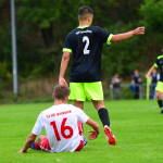Fußballer im schwarzen Trikot zeigt nach einem Spielzug Richtung, während ein anderer am Boden liegt; Schiedsrichter verfolgt das Geschehen.