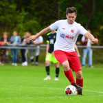 Fußballspieler im weißen Trikot und roten Hosen dribbelt auf dem Rasen, Blick auf den Ball; Hintergrund mit Zuschauern.