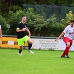 Drei Fußballspieler im Freien im Laufduell; einer lenkt den Ball mit dem Fuß, die anderen laufen mit, im Hintergrund Werbetafeln.