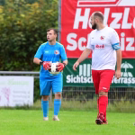 Zwei Fußballspieler im Lauf, einer in weiß-rot, der andere in blau, halten den Ball; Hintergrund mit rotem Sponsorbanner.