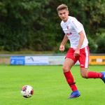 Fußballspieler in rotem Outfit dribbelt und schießt den Ball auf einem grünen Rasen, unscharfer Hintergrund im Stadion.