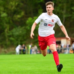 Fußballspieler im weißen Trikot und roten Shorts sprintet auf dem Rasen, Arme in Bewegung, während Zuschauer im Hintergrund zuschauen.