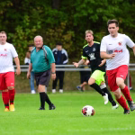 Fußballspiel im Freien: Spieler im Sprint kurz vorm Schuss; Schiedsrichter und weitere Akteure im Hintergrund, Rasen und Hecken.