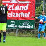 Fußballer im blauen Trikot hält einen Ball mit Handschuhen, während Gegenspieler im Vordergrund unscharf vorbeilaufen.