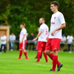 Fußballspieler in rotem Stutzen im Vordergrund, seitlich blickend, während im Hintergrund weitere Spieler unscharf spielen.
