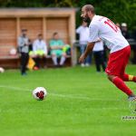 Fußballspieler im weißen Trikot mit rotem Hosenbein am Ball, kurz vor dem Schuss im Stadion; Spielszene im Fokus.