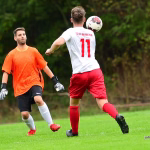 Zwei Fußballspieler im Spiel: einer wirft sich dem Ball entgegen, der andere läuft seitlich, vor unscharfem Stadionhintergrund.