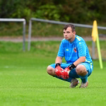 Fußballspieler kniet im Gras im Torraum und hält die Handschuhe im Einsatz, um den Ball abzuwehren.