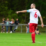 Fußballspieler in weißem Trikot und roten Hosen mit erhobenem Arm, läuft seitlich auf dem Spielfeld; Hintergrund unscharf.