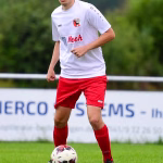 Fußballspieler im weißen Trikot und roten Hosen spielt den Ball im Lauf auf dem Rasen, im Hintergrund unscharfes Stadionpublikum.
