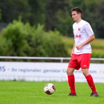 Fußballspieler im Aufwärmstil mit Ball am Boden, steht seitlich zum Spielgeschehen auf dem grünen Rasen.