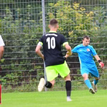 Fußballspiel: Spieler in Blau dribbelt am Ball vorbei, während ein Gegenspieler mit der Rückennummer 10 sprintet, vor einem Gitterzaun.