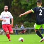 Fußballspiel: Spieler in Weiß-Rot und ein Gegenspieler in Schwarz-Neon laufen dem Ball nach, Szene einer Spielsituation am Sportplatz.