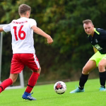 Zwei Fußballspieler im Spiel: Einer in Rot-Weiß mit Rückennummer, einer in Schwarz-Grün versucht den Ball zu stoppen auf dem Rasen.