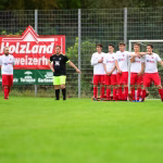 Fußballspieler in zwei Trikots stehen vor der Bande, eine Mannschaft in Weiß und Rot im Team-Display, im Hintergrund Zuschauerkulisse am Spielfeldrand.
