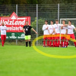 Fußballspieler stehen im Stadion neben einem Ball; eine Mannschaft in weiß-rot präsentiert sich, im Hintergrund Werbebanner und Tor.