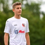 Sportler in weißem Trikot im Freien, Halbprofil mit Blick zur Seite vor unscharfem Grün-Hintergrund.