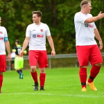 Fußballspieler in weißen Trikots und roten Hosen stehen im Trainingsfeld, ein Spieler winkt und zeigt, während andere zuschauen.