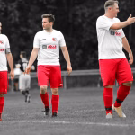 Drei Fußballspieler in weißen Trikots mit roten Hosen laufen im Stadion, einer gestikuliert zur Mannschaft; Spielszene vor dem Anstoß.