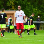 Fußballspieler im weißen Trikot mit roter Hose geht auf dem Rasen, während Gegenspieler im Vordergrund zurückblicken.