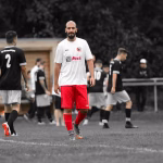 Fußballspieler in weißem Trikot und roten Hosen steht auf dem Platz, blickt nach vorn; im Hintergrund weitere Spieler während des Spiels.