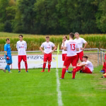 Fußballspiel: mehrere Spieler in roten Trikots stehen auf nassem Rasen, während ein Trainer am Spielfeldrand Anweisungen gibt.
