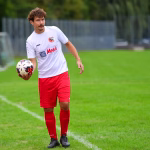 Fußballspieler in weißem Trikot und roten Hosen hält einen Ball in der Hand auf einem Rasenfeld; Sportaktion im Spielbetrieb.